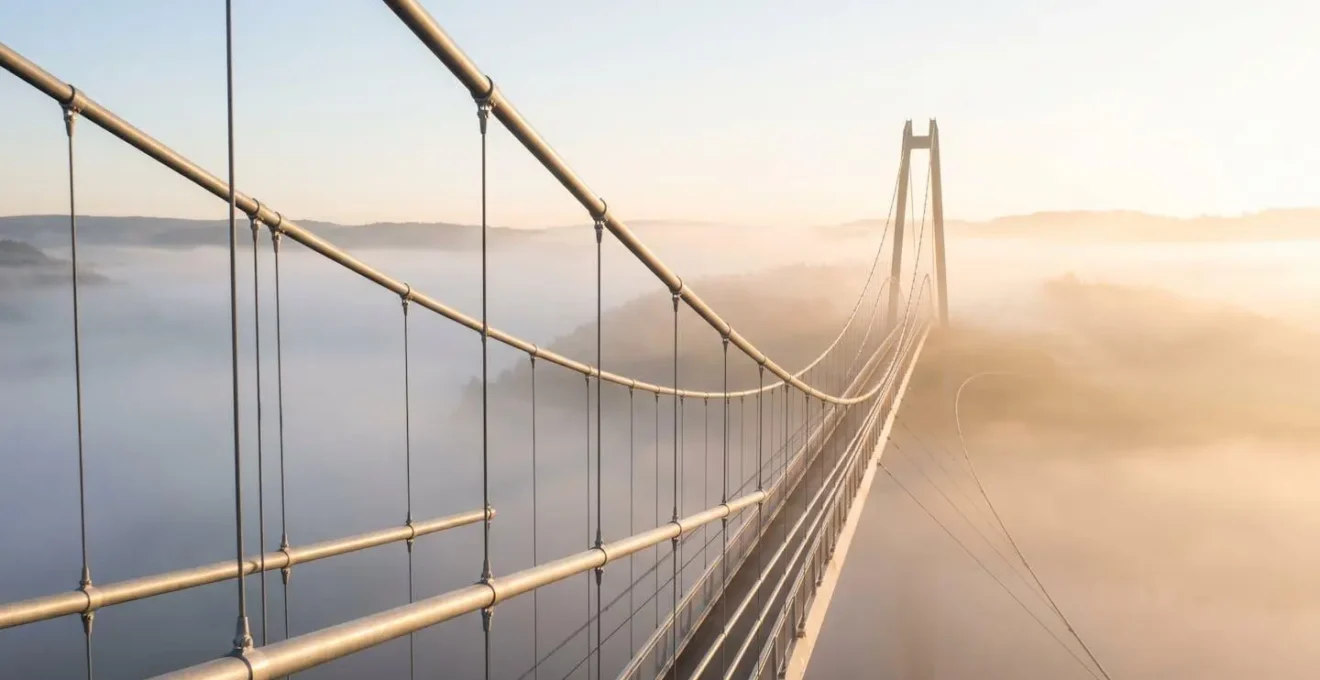 Pont suspendu moderne traversant une vallée brumeuse au lever du soleil avec lumière dorée sur les câbles métalliques